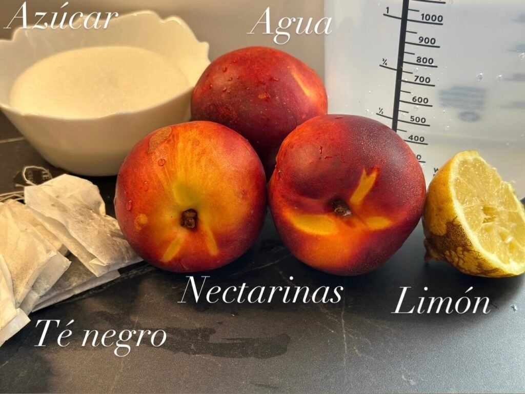 ingredientes en una encimera negra, tres nectarinas, medio limón, un vaso medidor con agua, bolsas de te negro y un cuenco con azúcar